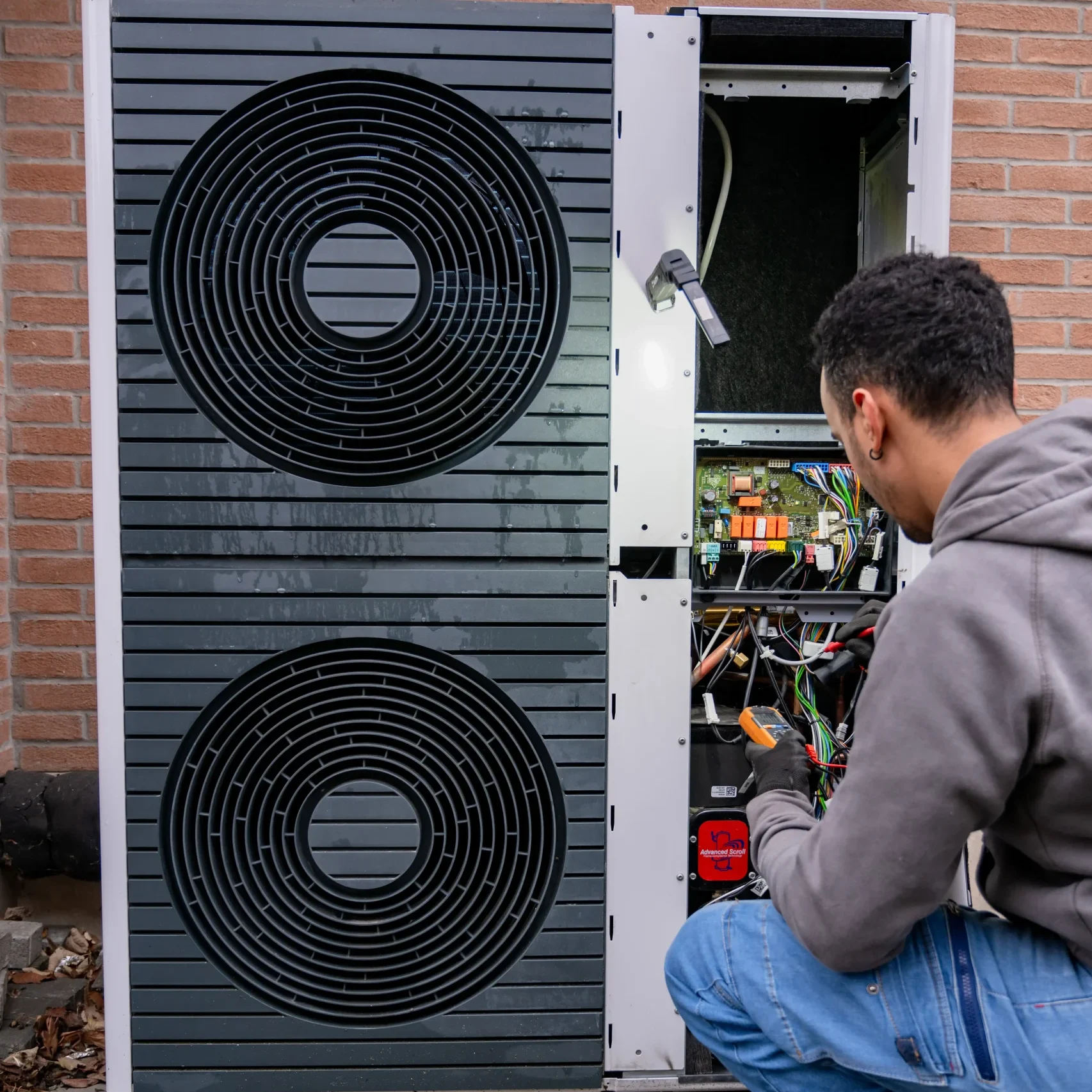 Un tecnico è inginocchiato davanti a un'unità esterna di climatizzazione o pompa di calore con la copertura aperta, esponendo i componenti interni. L'unità ha due grandi ventole nere visibili sul lato sinistro. Il tecnico, ,indossando guanti, sta controllando o riparando i cavi e i circuiti all'interno dell'unità. L'ambiente sembra esterno, con una parete di mattoni sullo sfondo. L'immagine trasmette l'attenzione al dettaglio e la cura nella manutenzione di sistemi di raffreddamento o riscaldamento complessi.