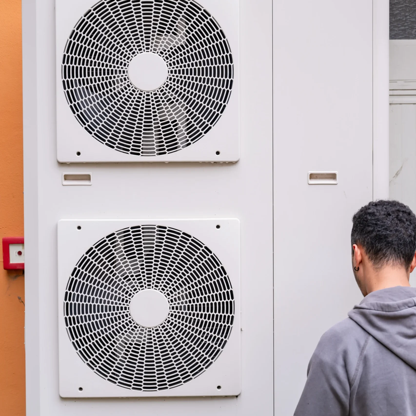 Un tecnico è davanti a un'unità esterna di climatizzazione o pompa di calore, composta da due grandi ventole bianche montate verticalmente. L'unità è installata su una parete esterna. L'immagine cattura un momento di lavoro o ispezione, trasmettendo un senso di professionalità e attenzione al dettaglio.