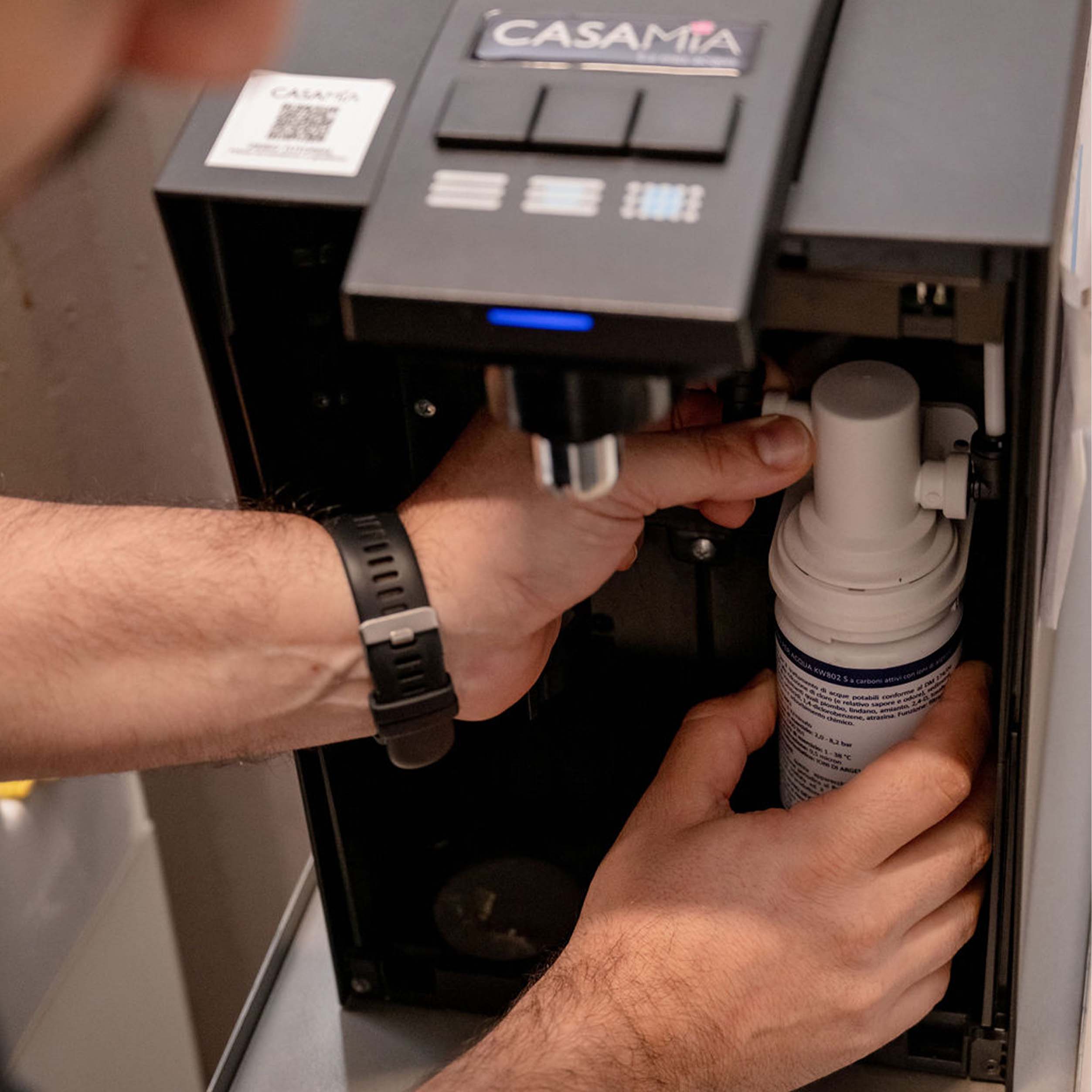 Tecnico interagisce con un distributore d'acqua. Sopra ai pulsanti, c'è il logo "CASAMIA". La persona ha le mani vicine al distributore, probabilmente per inserire o rimuovere un contenitore.
