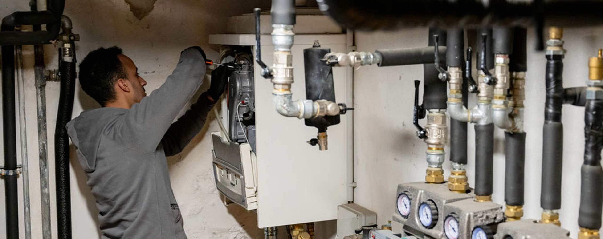 Un tecnico è intento a lavorare su un sistema di riscaldamento, eseguendo manutenzione su una caldaia. Indossa guanti e utilizza un cacciavite per regolare i componenti interni. Di fronte a lui, una complessa rete di tubazioni e valvole, alcune delle quali sono dotate di manometri per la lettura della pressione. Le tubature sono ben organizzate e mostrano segni di isolamento termico, tipico degli impianti di riscaldamento. L’ambiente ha un aspetto tecnico e funzionale, indicativo di una sala macchine o di un locale caldaia.